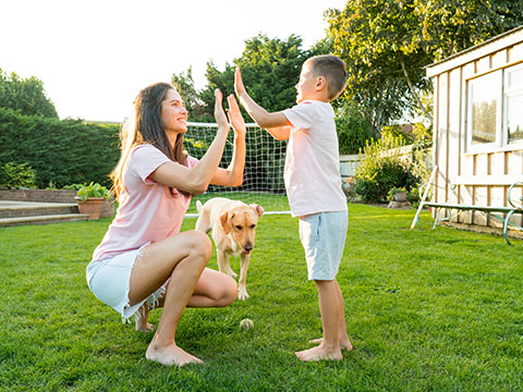 Young mother and son playing with dog, throwing a ball and have fun together. Happy family playing with tennis ball with pet. Fun Playing Games in Backyard Lawn on Sunny Summer Day. Active childhood.